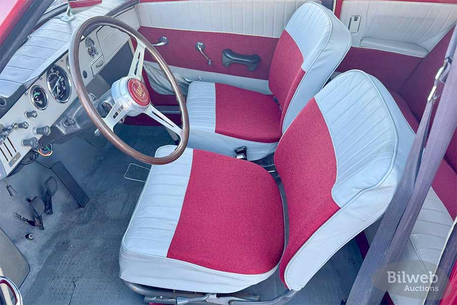 The interior of this 1964 Saab 96 2-Stroke remains in remarkable condition, featuring two-tone red and light gray upholstery that complements its rally heritage. The sporty four-bolt wooden steering wheel, a rare period-correct detail, adds to the driver-focused experience.