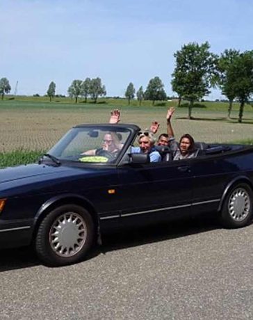 Joyful participants waving from a classic Saab convertible on a sunny day during the Roll on Roll off charity drive event