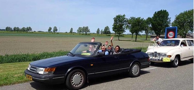 Joyful participants waving from a classic Saab convertible on a sunny day during the Roll on Roll off charity drive event