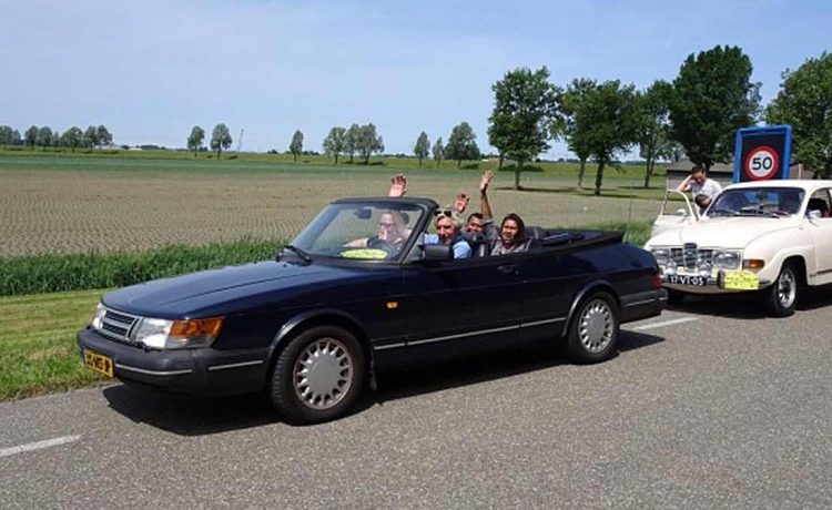 Joyful participants waving from a classic Saab convertible on a sunny day during the Roll on Roll off charity drive event