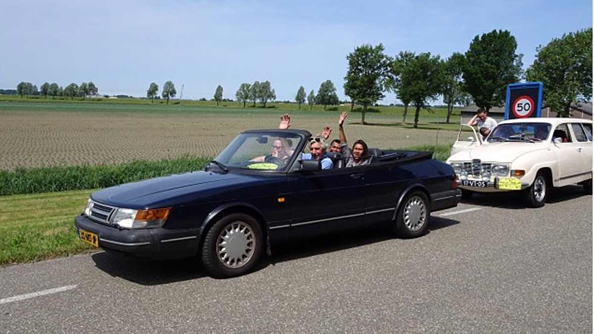 Joyful participants waving from a classic Saab convertible on a sunny day during the Roll on Roll off charity drive event