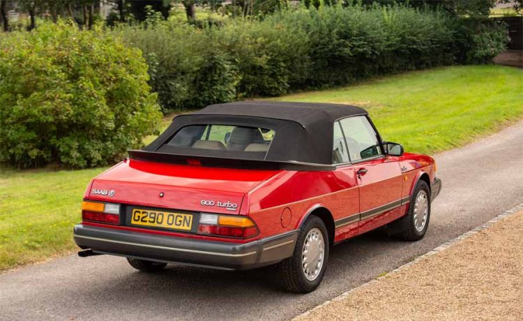 the iconic 1990 Saab 900 Turbo 16 Convertible in vibrant red, a classic symbol of sophistication and performance, as it stands gracefully in a lush setting.