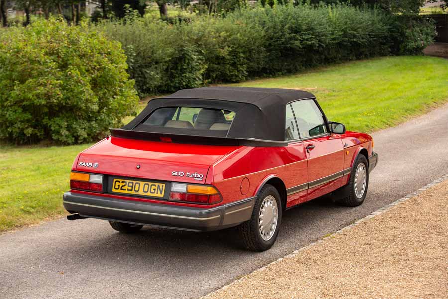 the iconic 1990 Saab 900 Turbo 16 Convertible in vibrant red, a classic symbol of sophistication and performance, as it stands gracefully in a lush setting.