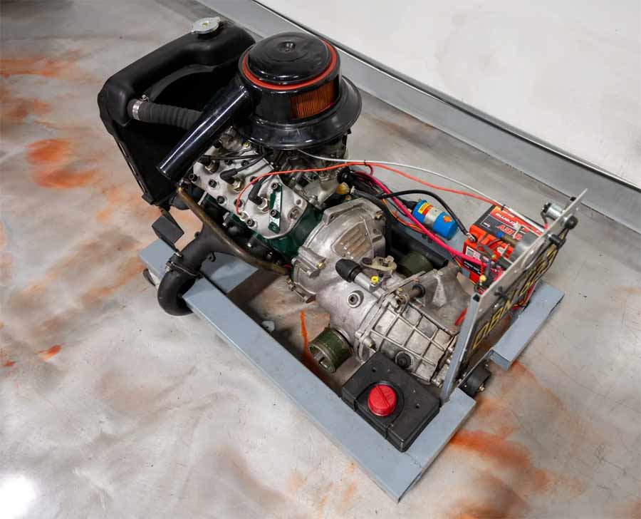 Overhead view of a meticulously assembled Saab 841cc two-stroke engine and transmission system mounted on a mobile test stand, featuring an array of neatly arranged components including carburetors, an air cleaner, ignition system, and a control panel with a red emergency stop button, all set against a workshop floor.