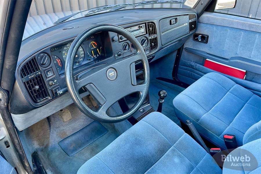 The image displays the interior of the classic 1983 Saab 900 Turbo APC, featuring its functional and driver-oriented cockpit. The blue fabric seats are in remarkable condition, echoing the vehicle’s era with their firm structure and distinctive texture. The instrument cluster remains fully intact, with original gauges that monitor the heart of the Turbo APC system.