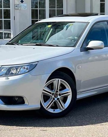 An exquisite Snow Silver Saab 9-3 SportEstate Turbo X with a panoramic sunroof parked outside a modern building, highlighting its sleek design and advanced engineering for the discerning collector.