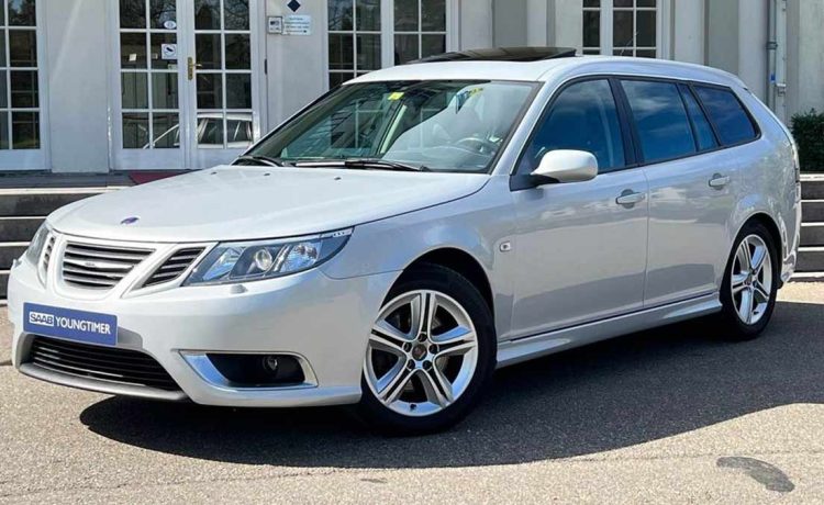 An exquisite Snow Silver Saab 9-3 SportEstate Turbo X with a panoramic sunroof parked outside a modern building, highlighting its sleek design and advanced engineering for the discerning collector.