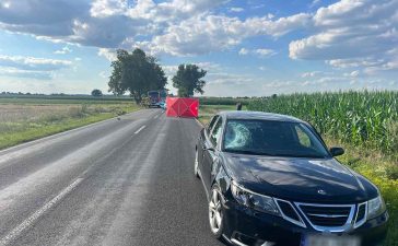 The image captures the aftermath of a tragic accident in Gołąbki, showing a Saab vehicle with a damaged windshield, indicative of the impact with the cyclist. Emergency services and police are present at the scene, conducting their investigation.