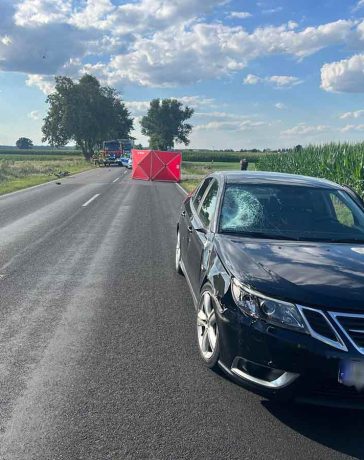 The image captures the aftermath of a tragic accident in Gołąbki, showing a Saab vehicle with a damaged windshield, indicative of the impact with the cyclist. Emergency services and police are present at the scene, conducting their investigation.