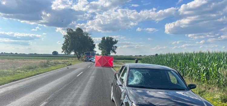 The image captures the aftermath of a tragic accident in Gołąbki, showing a Saab vehicle with a damaged windshield, indicative of the impact with the cyclist. Emergency services and police are present at the scene, conducting their investigation.