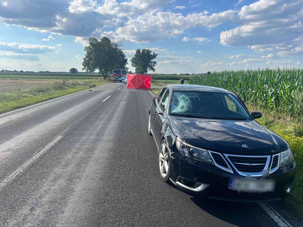 The image captures the aftermath of a tragic accident in Gołąbki, showing a Saab vehicle with a damaged windshield, indicative of the impact with the cyclist. Emergency services and police are present at the scene, conducting their investigation.