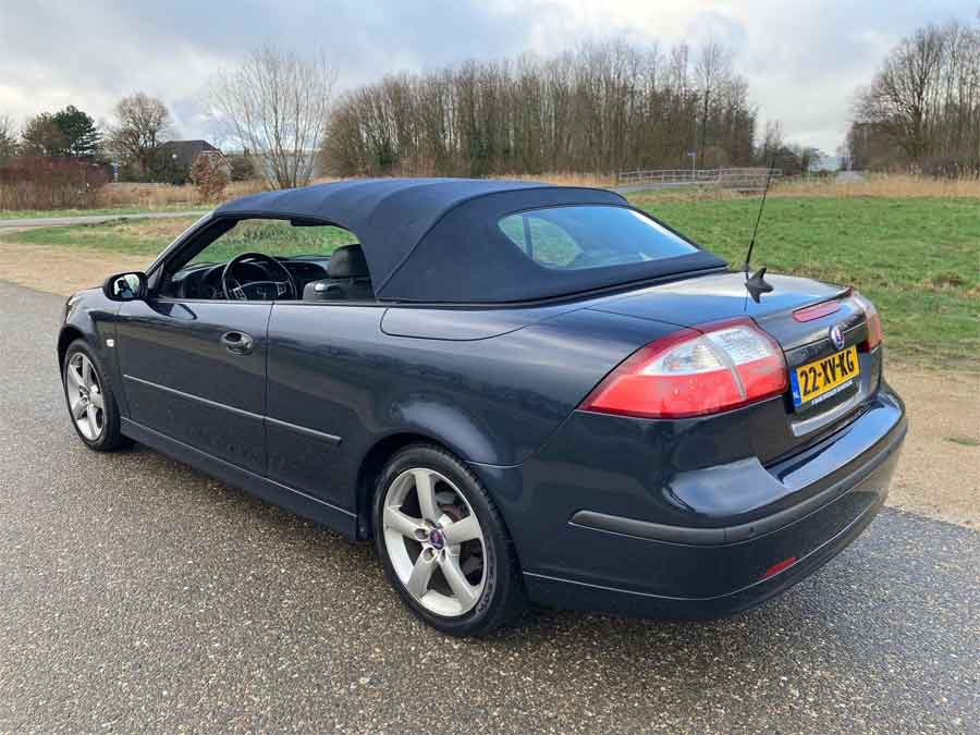 A view of a dark blue Saab 9-3 Vector Cabriolet with the soft top up, highlighting its sleek design and sporty profile, set against a serene outdoor backdrop.