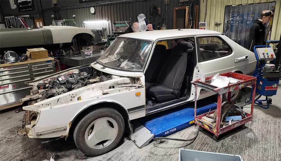 a Saab 900i serving as a donor car for a future restomod project. The car is positioned in a workshop, with various tools and equipment visible around it. The front end of the car has been disassembled, exposing the engine bay and chassis, indicating that it is in the initial stages of modification. The interior appears mostly intact, with seats still in place. This Saab 900i will undergo significant transformations as it contributes parts and structure to the ambitious restomod project, blending classic design with modern performance enhancements.