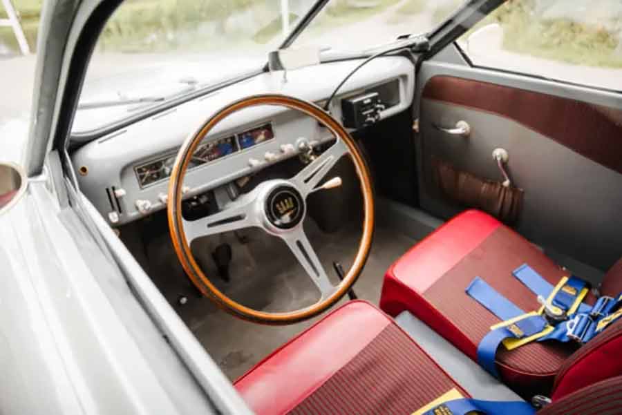 The meticulously restored interior of the 1957 Saab 93A—featuring red vinyl bucket seats with striped cloth inserts, a Saab Sport wood-rimmed steering wheel, and a Halda Tripmaster—fully equipped for rally adventures.