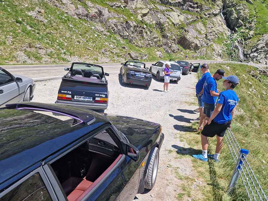 Saab enthusiasts take a break during their Transylvania tour, enjoying the stunning mountain scenery and camaraderie among fellow car lovers.