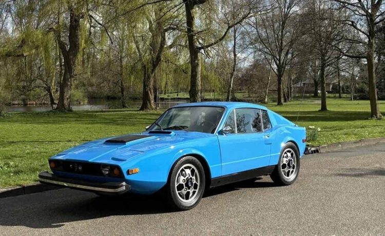 A vibrant 1974 Saab Sonett III in True Blue, showcasing its sleek design and sporty Soccerball wheels, poised in a serene park setting.