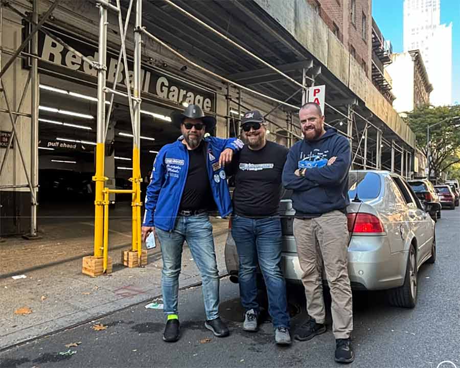 The record-breaking trio—Christopher Michaels, Nik Krueger, and Wes Vigh—stand proudly with their Saab 9-5 Aero at the iconic Red Ball Garage after conquering the Cannonball Run.