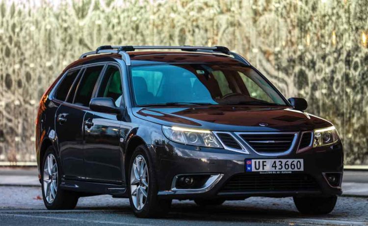 A sleek 2009 Saab 9-3 2.0T XWD Aut SportCombi Aero in Carbon Grey Metallic, parked against a serene backdrop showcasing Norway's stunning natural beauty, with the majestic mountains of Tromsø in the distance. The car's elegant matte chrome roof rails gleam under the soft northern sunlight, while its 17-inch aluminum wheels and the subtle sophistication of its design invite admiration.