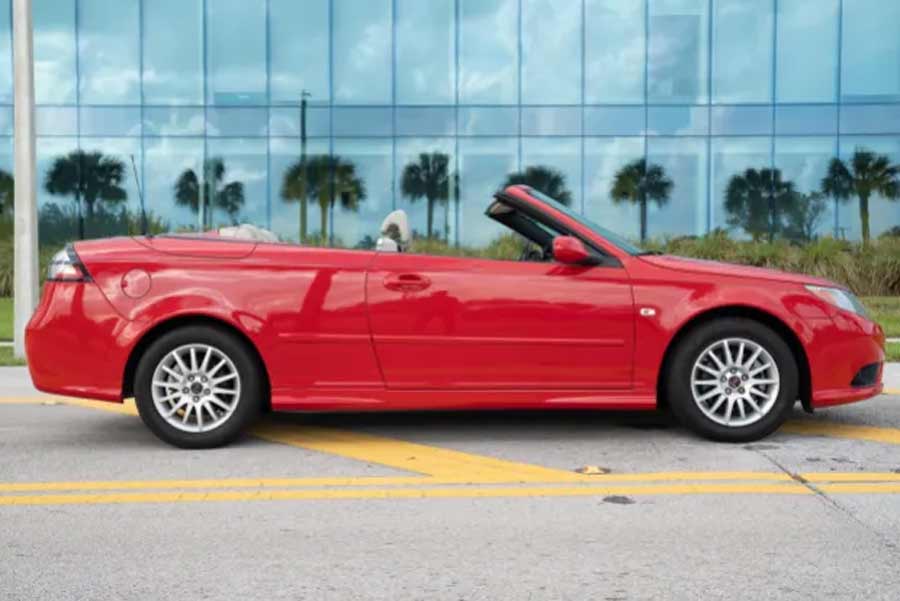 A gleaming 2008 Saab 9-3 Convertible in vibrant Laser Red, capturing the essence of Florida's charm with palm trees mirrored in the sleek glass facade of a nearby building