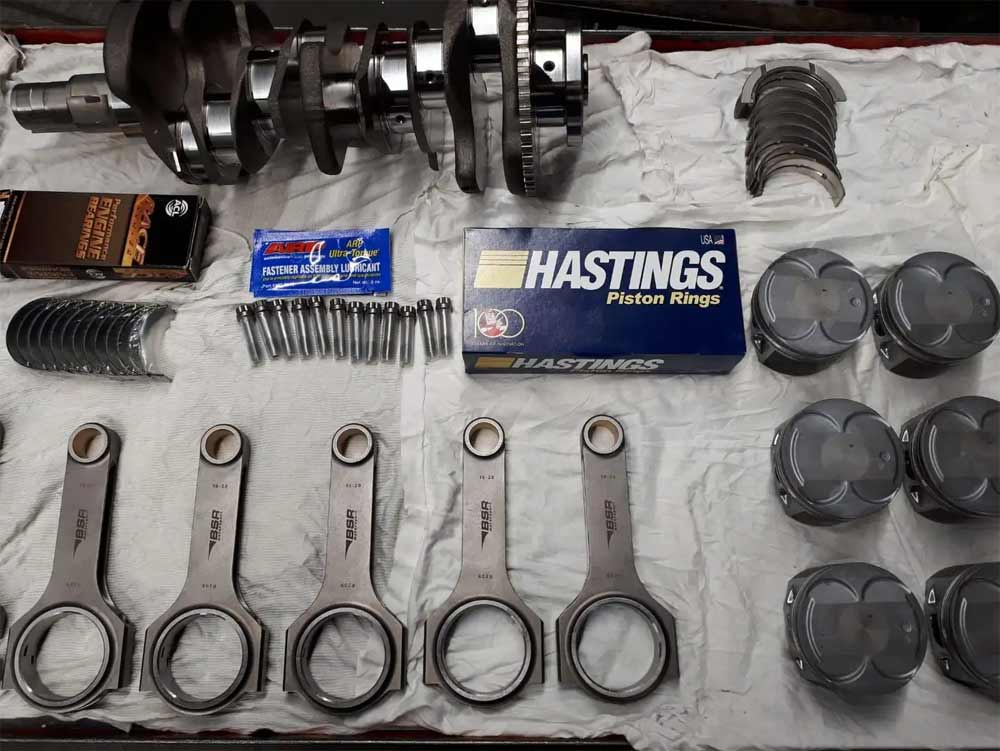 High-performance tuning components laid out for a Saab 2.8 engine upgrade, including a crankshaft, Hastings piston rings, ARP fastener assembly lubricant, connecting rods, and pistons, ready for assembly to boost engine power and efficiency.