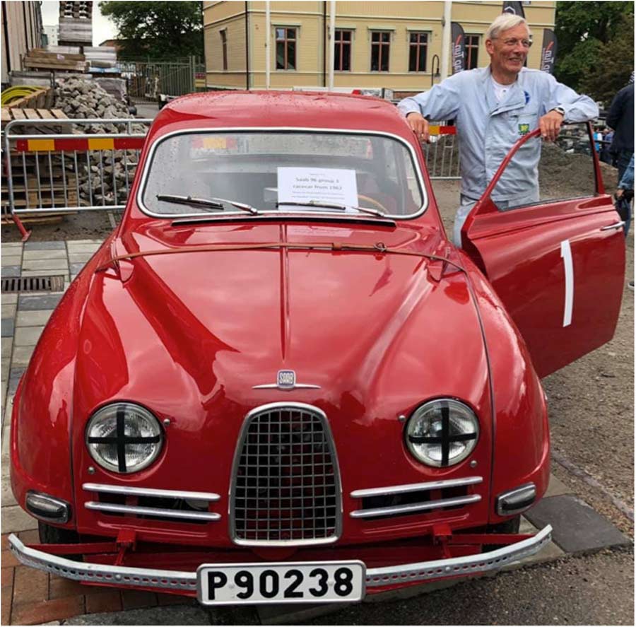 Ulf G Andersson stands beside the Group 3 Saab rally car at an automotive festival, donning Gösta Karlsson's racing jumpsuit