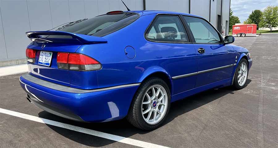  the 1999 Saab 9-3 Viggen in Lightning Blue Metallic, highlighting its distinctive rear spoiler, sleek body lines, and sporty 17" BBS wheels. The car's vibrant color and aerodynamic design features, including the rocker panel extensions, enhance its dynamic appearance.