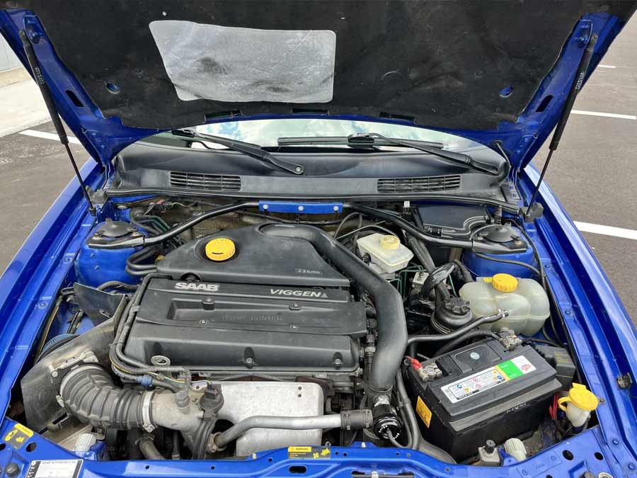 The engine bay of the 1999 Saab 9-3 Viggen, housing a turbocharged 2.3-liter inline-four engine. This replacement engine, sourced from a 2001 Saab 9-3 Viggen, delivers impressive performance and reliability. Key features include the Saab Viggen-branded engine cover, a replacement intake, and a Forge 007 bypass valve. The engine bay is well-maintained, reflecting the care taken by its owner to ensure optimal performance.